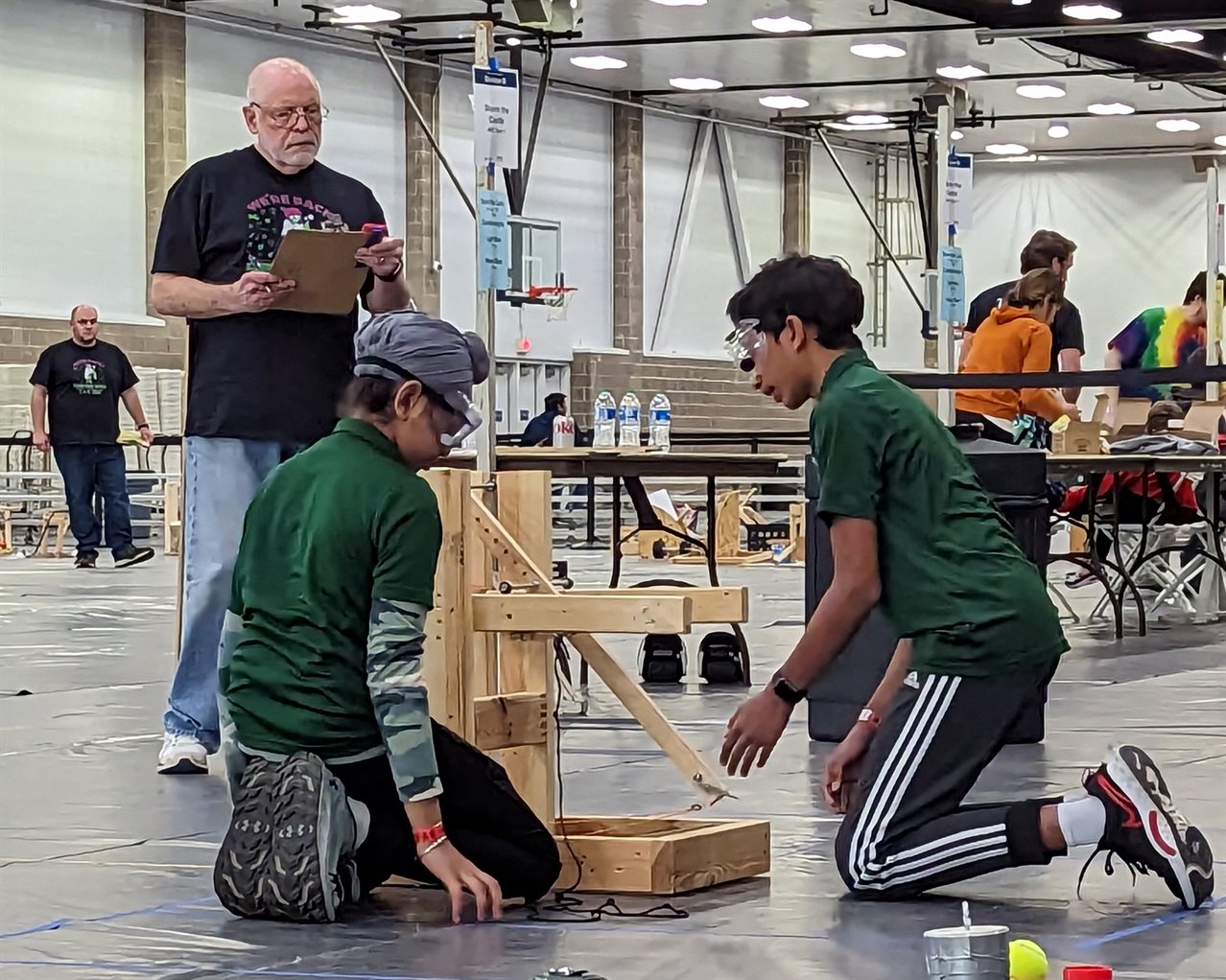 2 Young students working on a STEM challenge project at the 2023 Illinois Science Olympiad while a judge watches.